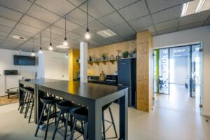 Moderne kantoorkeuken aan de De Ruijterkade met hoge tafels, barkrukken en een open doorgang naar een vergaderruimte.